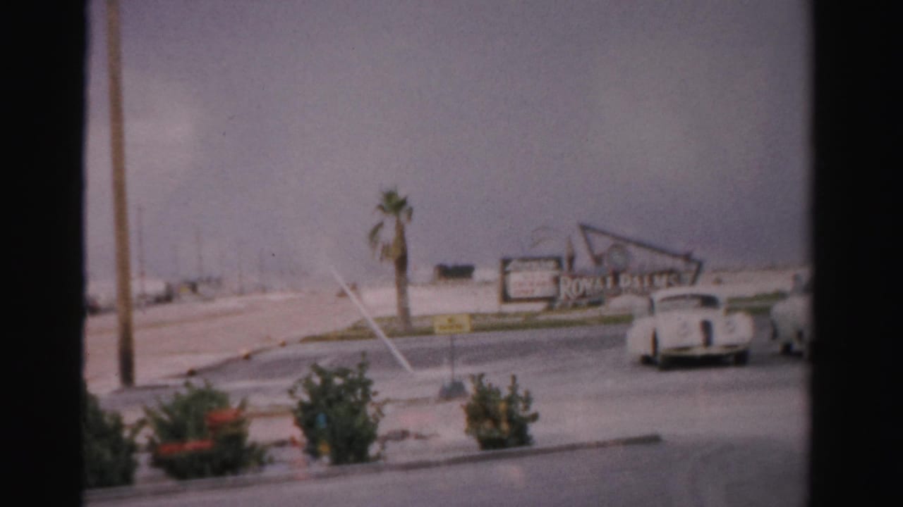 1957 Las Vegas home-movie frame showing classic cars on a wide road near palms, shrubs, and a roadside sign.