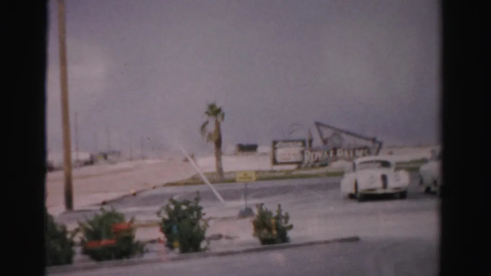 1957 Las Vegas home-movie frame showing classic cars on a wide road near palms, shrubs, and a roadside sign.