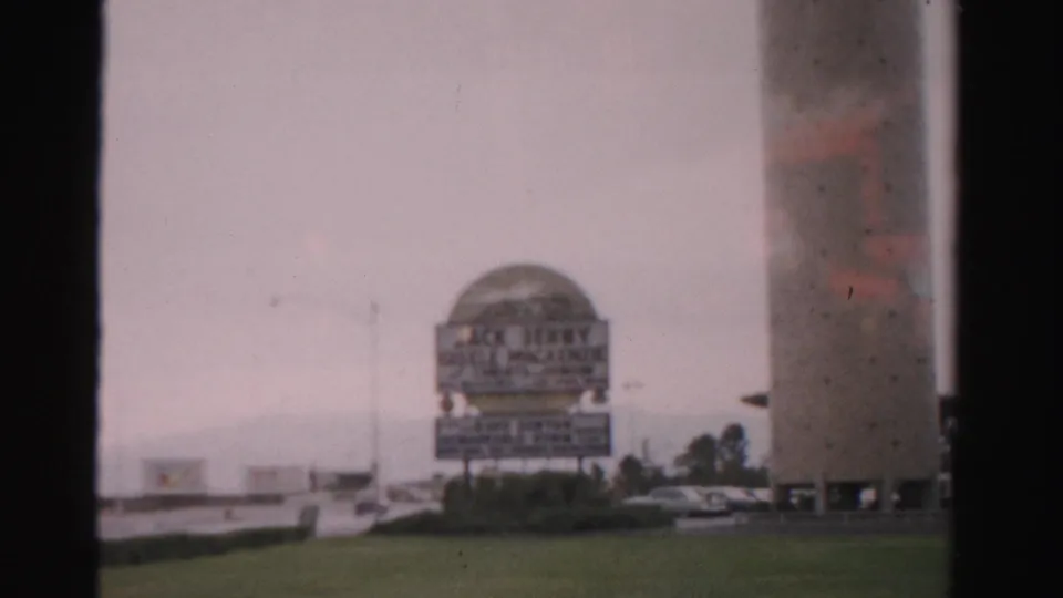 1957 Las Vegas frame with a dome-topped sign structure, tall cylindrical building, lawn, cars, and palms.