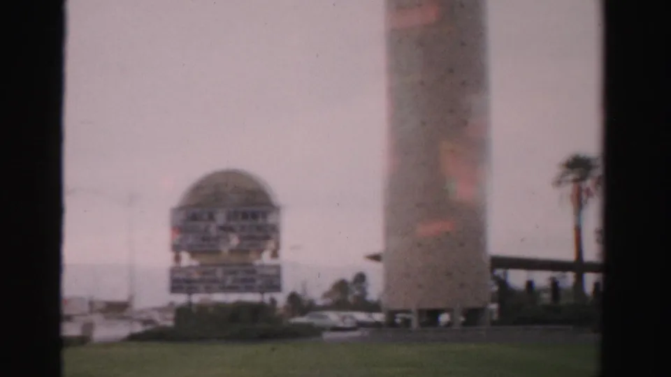 Soft 8mm Las Vegas frame showing a cylindrical building, dome-shaped sign, palm, lawn, and roadside structures.