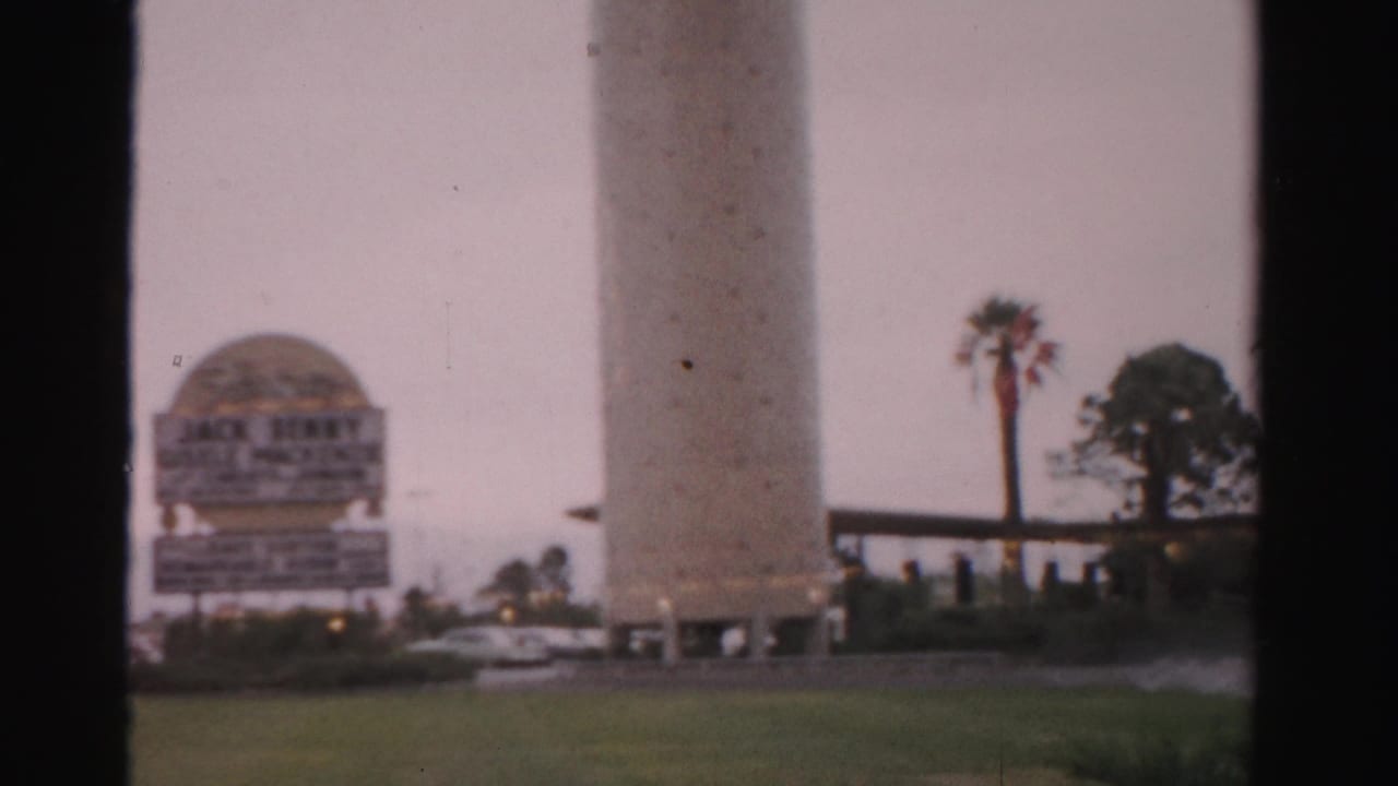 1957 Las Vegas home-movie frame showing a tall cylindrical tower, a domed roadside sign structure, palms, lawn, and visible 8mm frame edges.