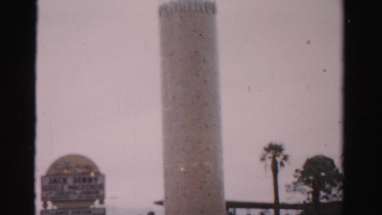 1957 Las Vegas 8mm frame with a tall tower, domed sign structure, palm trees, and dark film-frame borders interrupting the view.