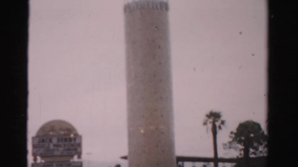 1957 Las Vegas 8mm frame with a tall tower, domed sign structure, palm trees, and dark film-frame borders interrupting the view.