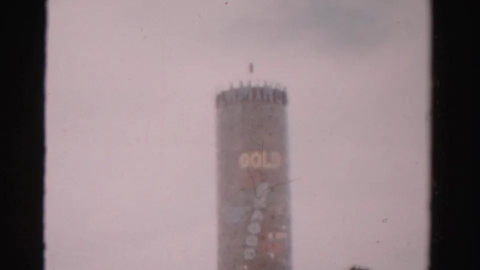 1957 Las Vegas 8mm frame showing the tall cylindrical tower with faint signage, pale overcast sky, film marks, and a palm near the lower edge.