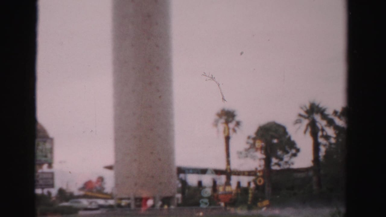1957 Las Vegas home-movie frame showing palm trees, roadside signage, and a hazy view through dark window or vehicle framing.