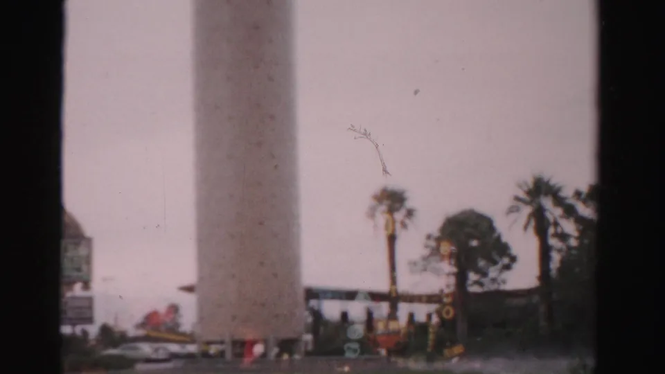 1957 Las Vegas home-movie frame showing palm trees, roadside signage, and a hazy view through dark window or vehicle framing.
