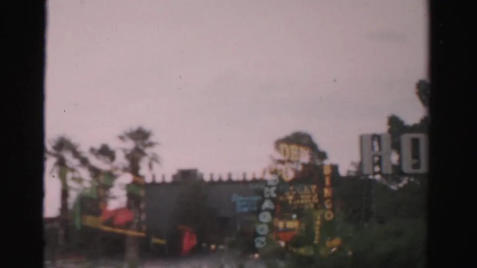1957 Las Vegas home-movie frame showing blurred hotel signage, palm trees, and low roadside buildings under grey sky.