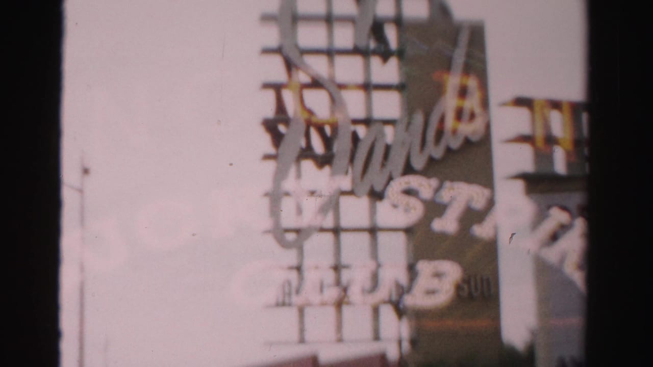 Soft 1957 Las Vegas 8mm frame showing a large exterior sign structure, partial lettering, street edge, and commercial building beyond.