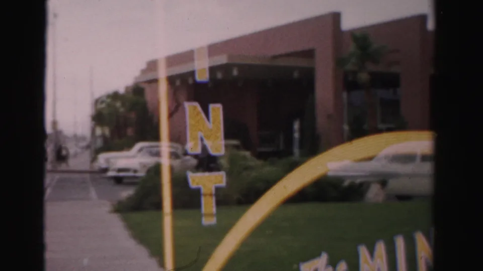 Visitor-shot 1957 Las Vegas frame with a sidewalk, lawn, yellow arch detail, mid-century cars, and commercial entrance.