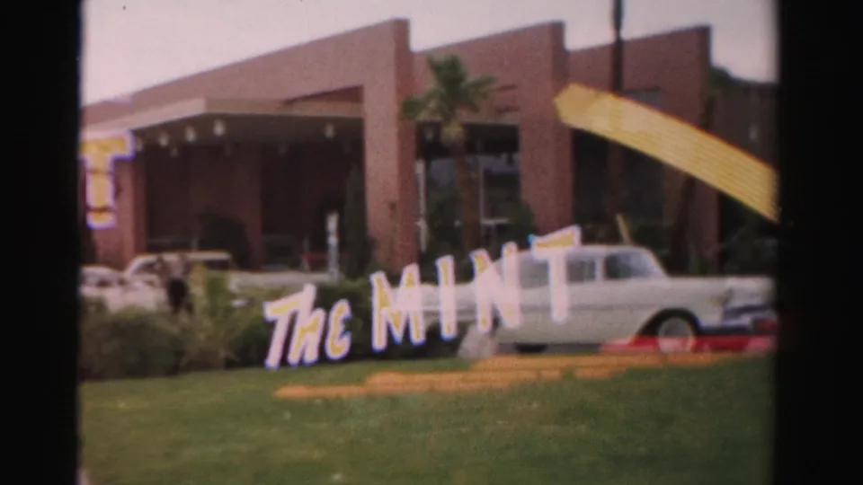1957 Las Vegas frame showing The Mint sign on a lawn, a white mid-century car, palms, shrubs, and a red-brown building.