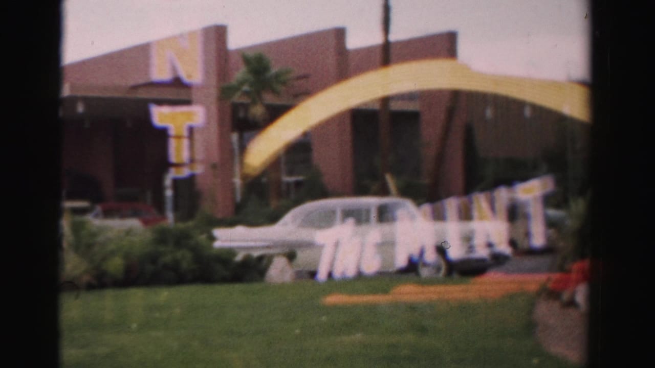 1957 Las Vegas home-movie frame showing The Mint sign elements, a white sedan, lawn, palm trees, and mid-century exterior facade.