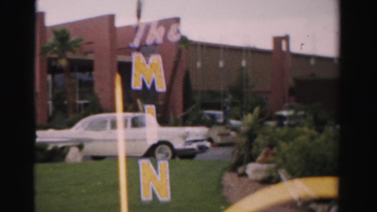 1957 Las Vegas frame with a white 1950s sedan in front of The Mint exterior and vertical yellow-blue sign lettering.
