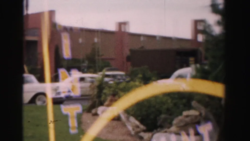 1957 Las Vegas home-movie view of partial The Mint signage, parked white car, yellow sign structure, rocks, plants, and commercial facade.