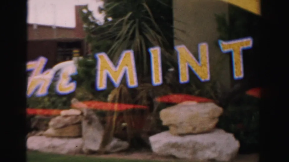 1957 Las Vegas frame showing large The Mint lettering with palm fronds, rock landscaping, red umbrellas, and parts of the building exterior.