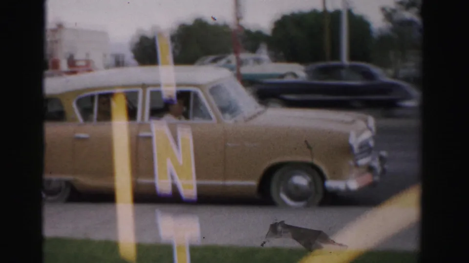 The beige station wagon continues past foreground The Mint lettering as another dark car moves along the Las Vegas street.