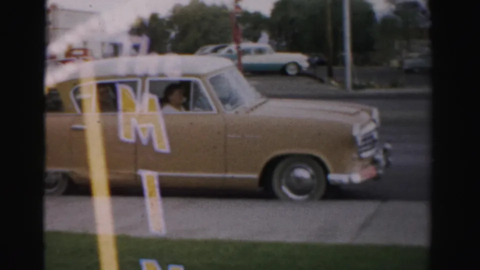 1957 Las Vegas street frame with a tan station wagon, white roof, The Mint lettering, grass verge, and a visible passenger face.