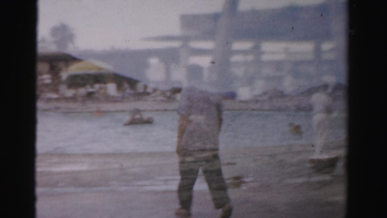 Man in a light shirt walks across wet sand near shallow water with beach structures in the distance.