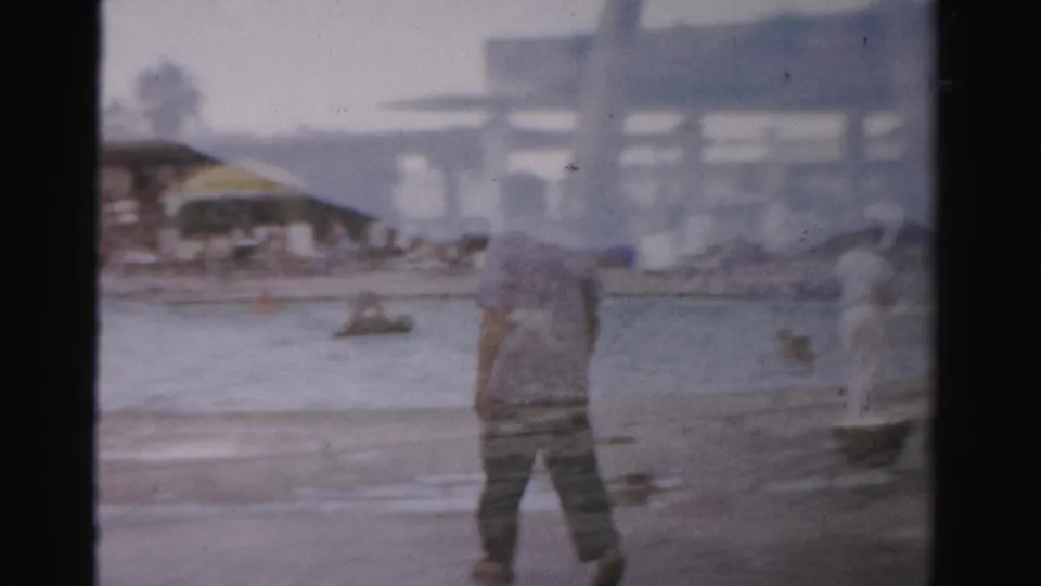 In 1957 Las Vegas-area home-movie footage, a man in a light shirt walks toward shallow water as distant swimmers and beach structures appear through haze.