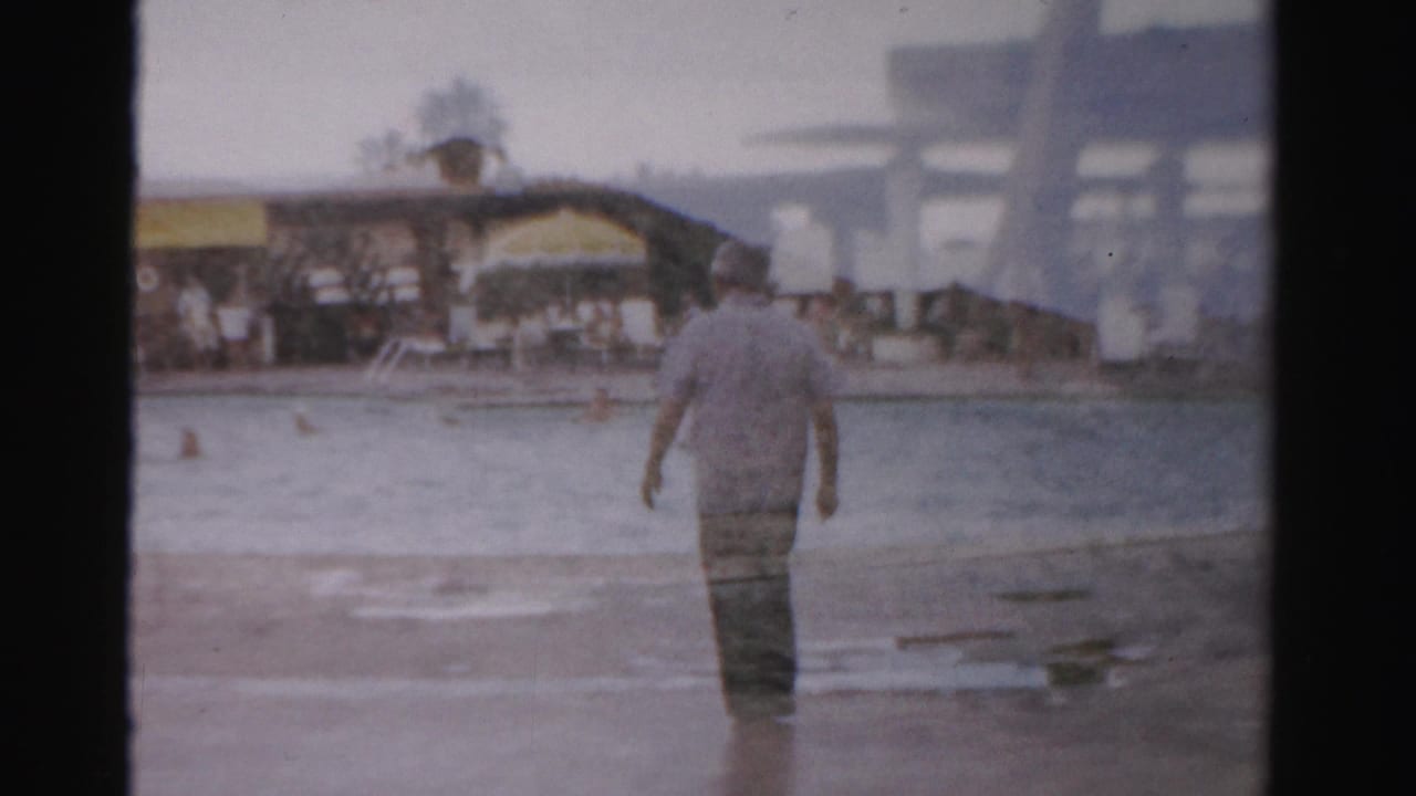 Man walks away from the camera through shallow water near a beach with swimmers and distant structures.