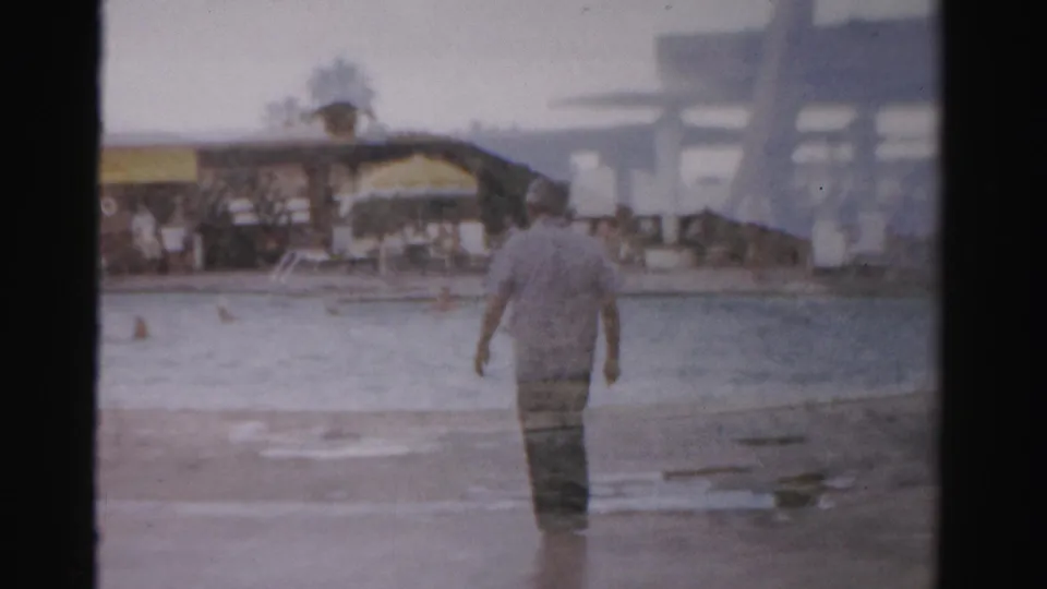 The man continues away from the camera in shallow water, with swimmers, a beach edge, and distant Las Vegas-area recreation structures behind him.