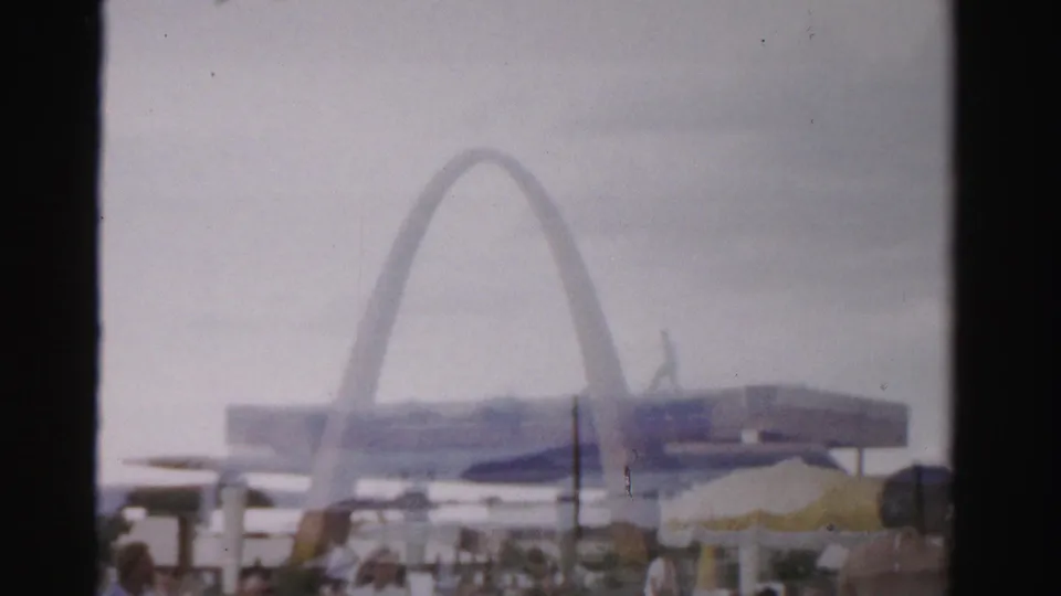 Faded 1957 Las Vegas frame with a large arched structure beyond outdoor tables, umbrellas, and distant visitors.