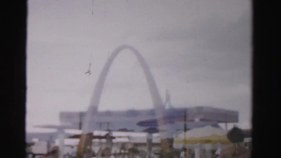 1957 Las Vegas home-movie frame showing a hazy pool area with bathers, umbrellas, low buildings, and a large arch-shaped structure in the background.