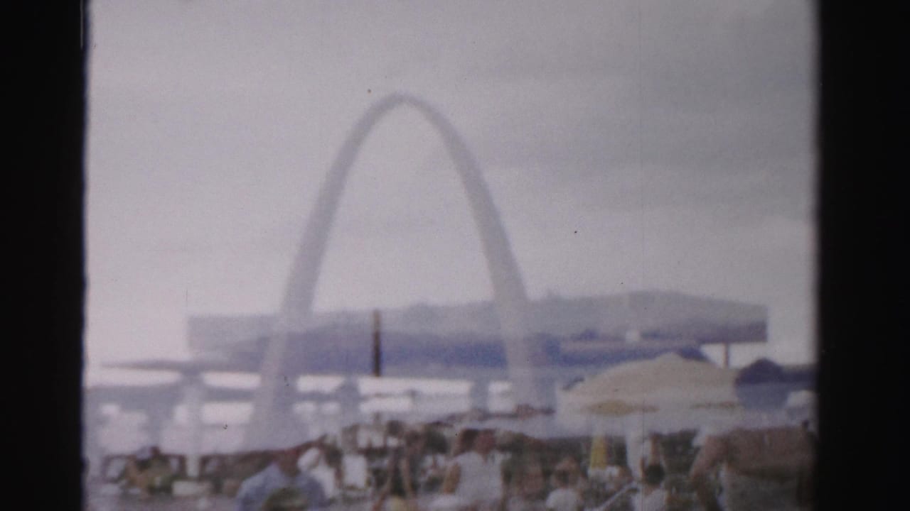 Poolside crowd under umbrellas with a large pale arch and buildings in the background.