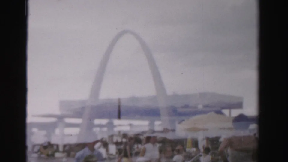 1957 Las Vegas resort pool view with people gathered under umbrellas, soft haze, and a large arch-shaped structure rising behind low buildings.