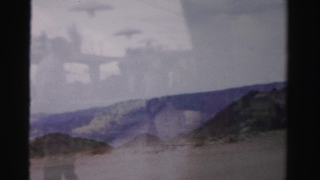 1957 Las Vegas home-movie frame showing a person walking near a roadside area with low rocks and soft, hazy exposure.