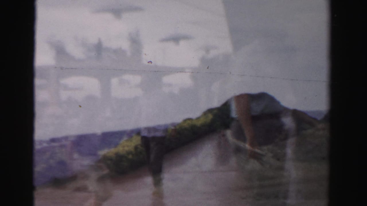 1957 Las Vegas frame with a person walking beside low landscaping or rocks, seen through soft 8mm focus and muted color.