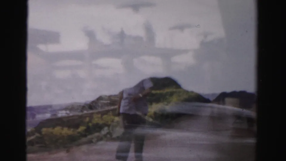 Home-movie frame from 1957 Las Vegas showing a person near a rocky roadside foreground with indistinct street structures behind.