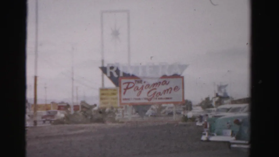 1957 Las Vegas street frame showing a large Pajama Game sign, mid-century cars, roadside poles, and hazy 8mm color.