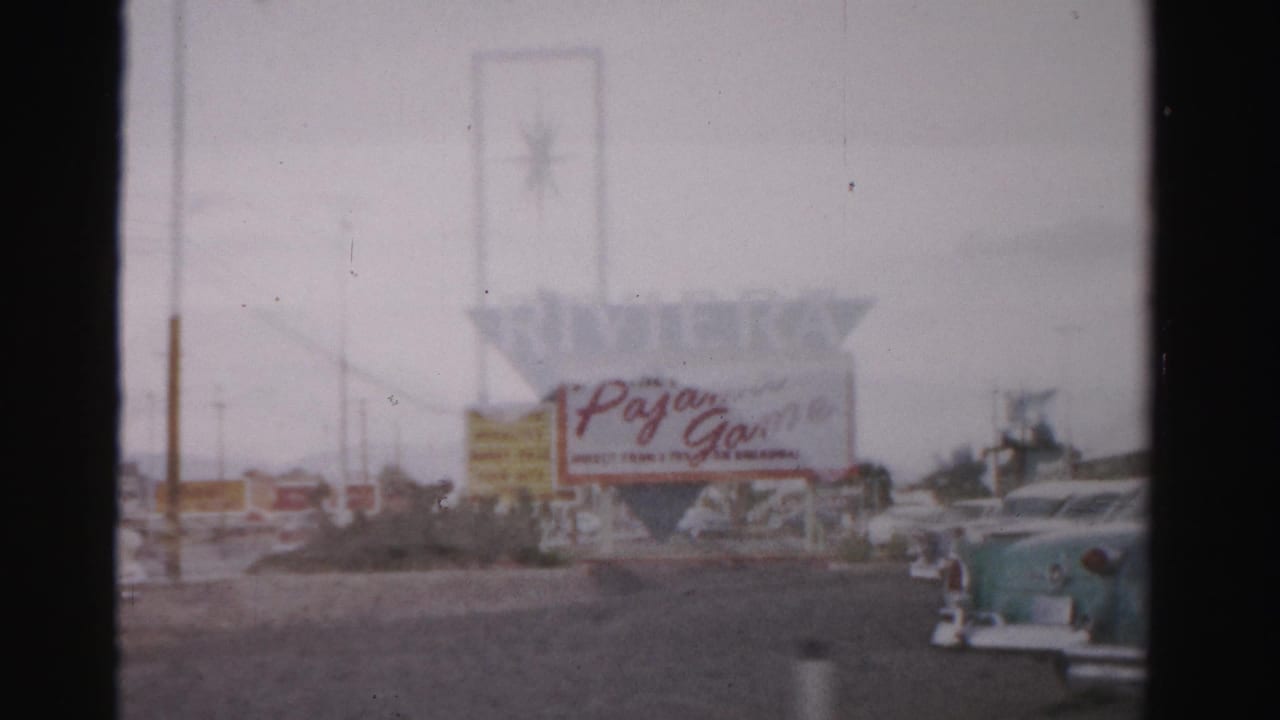 1957 Las Vegas frame showing the Riviera sign, Pajama Game marquee, roadside poles, and parked mid-century cars in soft 8mm color.