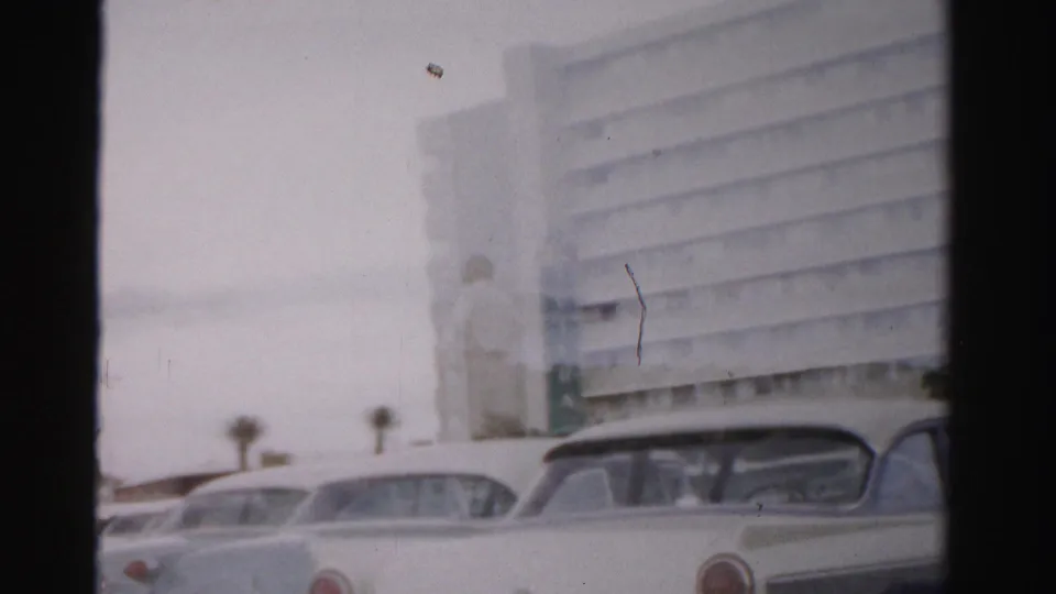 1957 Las Vegas frame showing a large hotel building, roadside sign area, palm trees, and a pale station wagon in soft home-movie color.