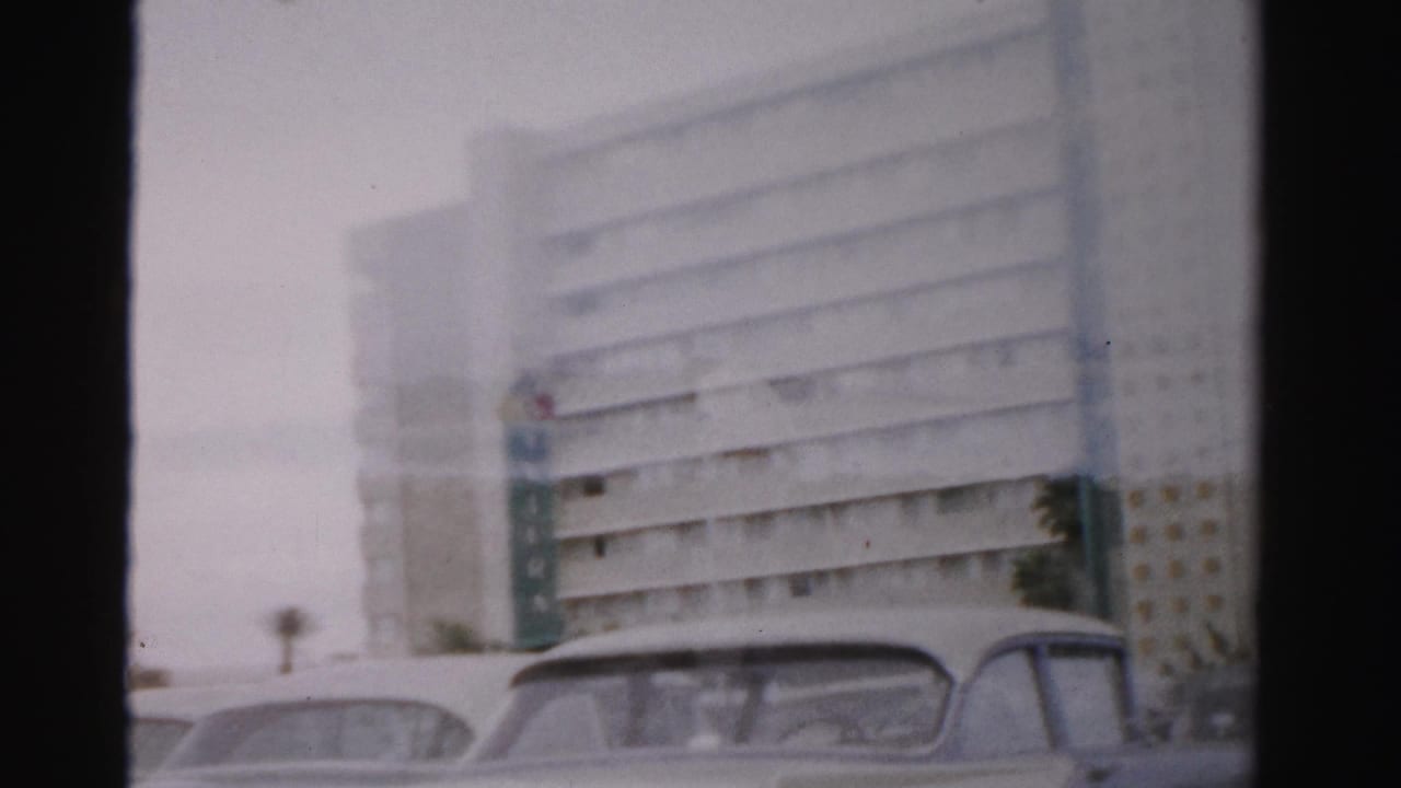 1957 Las Vegas home-movie frame showing a large modernist building and parked classic cars seen through soft 8mm haze.