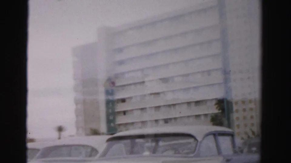 1957 Las Vegas home-movie frame showing a large modernist building and parked classic cars seen through soft 8mm haze.