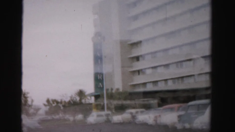 1957 Las Vegas 8mm view of a vertical sign near a large building, with classic cars parked along the roadside.