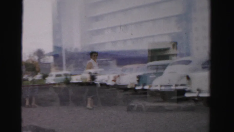 1957 Las Vegas frame showing a pedestrian by a parking area, classic cars, and a large roadside building in soft focus.
