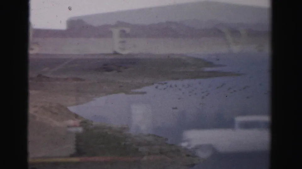 1957 Las Vegas home-movie frame showing a hazy lake or reservoir view with desert mountains and faint Royal Nevada sign lettering.