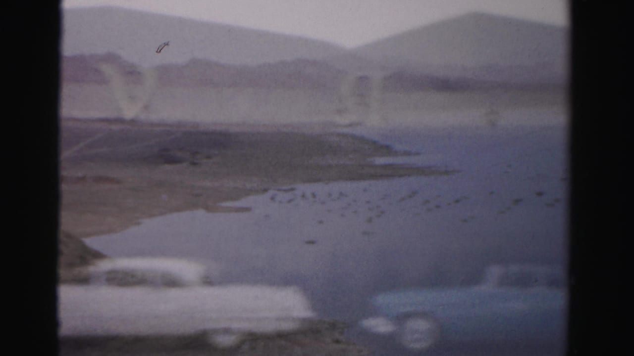 1957 Las Vegas 8mm frame with a reservoir-like view, distant hills, blurred parked cars, and visible Royal Nevada sign letters.