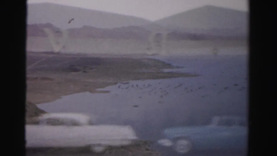 1957 Las Vegas 8mm frame with a reservoir-like view, distant hills, blurred parked cars, and visible Royal Nevada sign letters.