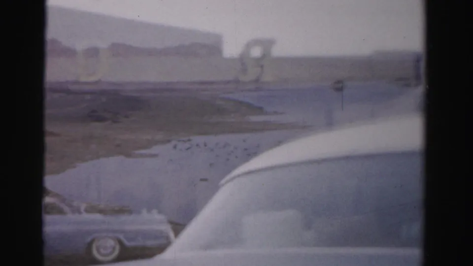1957 Las Vegas home-movie frame showing parked mid-century cars beside a lake or reservoir with mountains and partial Royal Nevada signage.