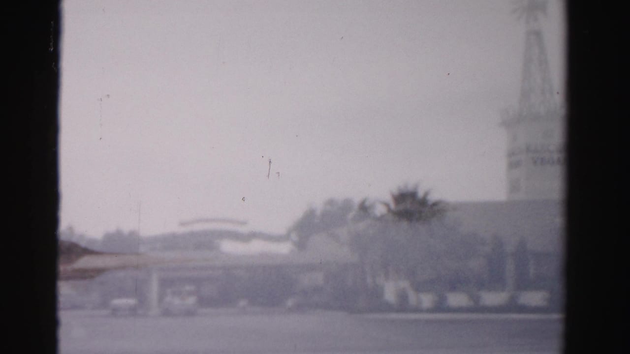 Soft view of a white windmill-style tower behind palms and low buildings under a cloudy sky.