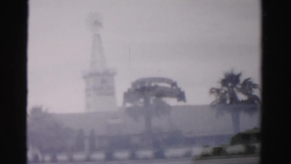 1957 Las Vegas home-movie frame showing the windmill-style tower, roadside palms, low structures, and partially visible cars.
