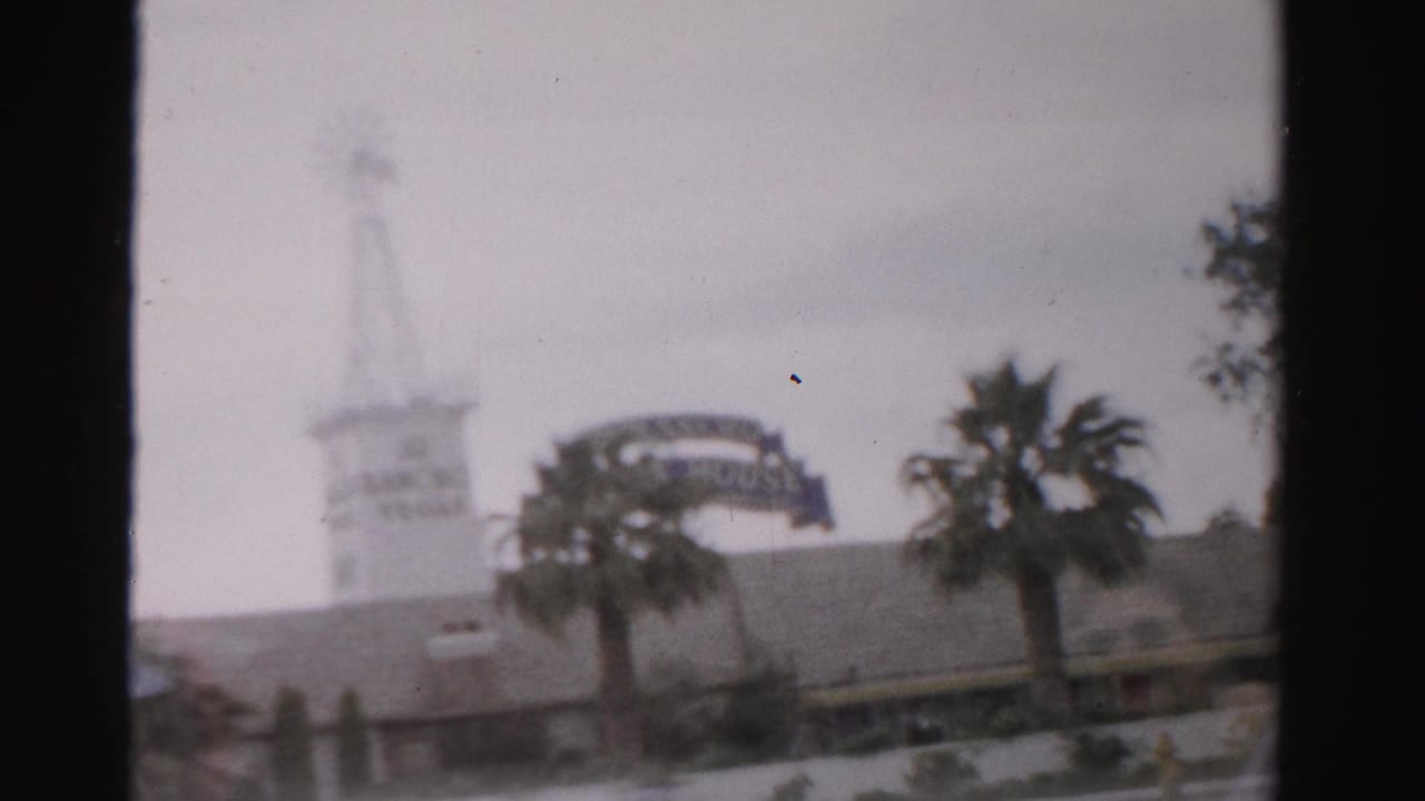1957 Las Vegas home-movie frame showing a hazy exterior with a windmill-like tower, curved sign structure, palm trees, and cloudy sky.