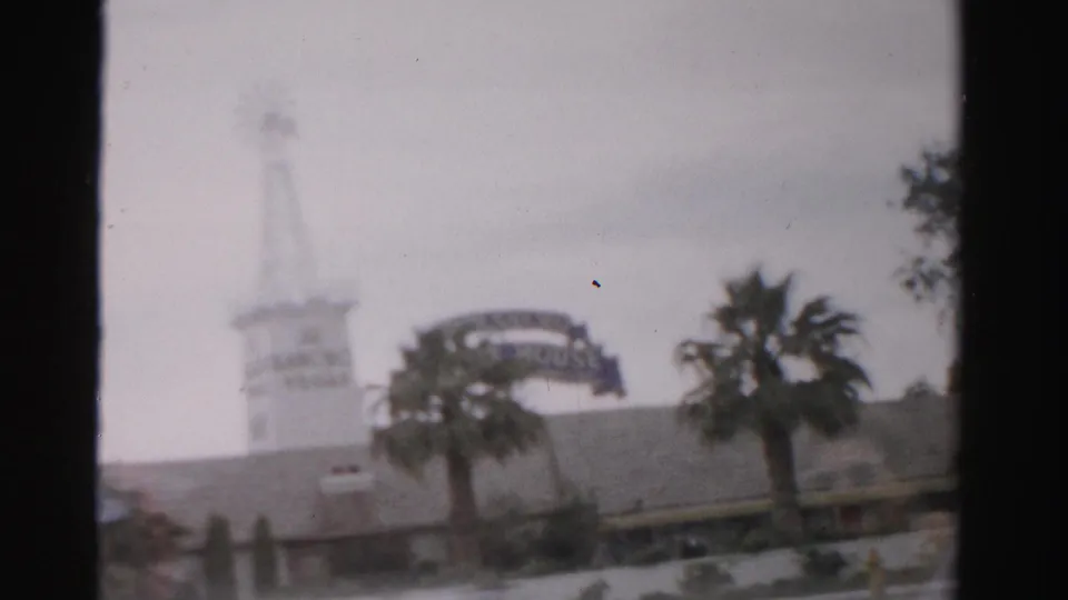 1957 Las Vegas home-movie frame showing a hazy exterior with a windmill-like tower, curved sign structure, palm trees, and cloudy sky.