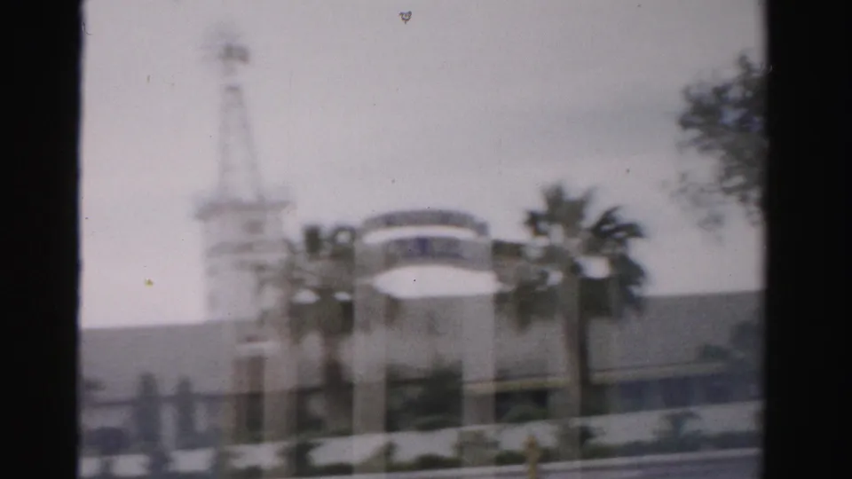 1957 Las Vegas home-movie view showing a blurred windmill-like tower, arched exterior feature, palms, and roadway edge.