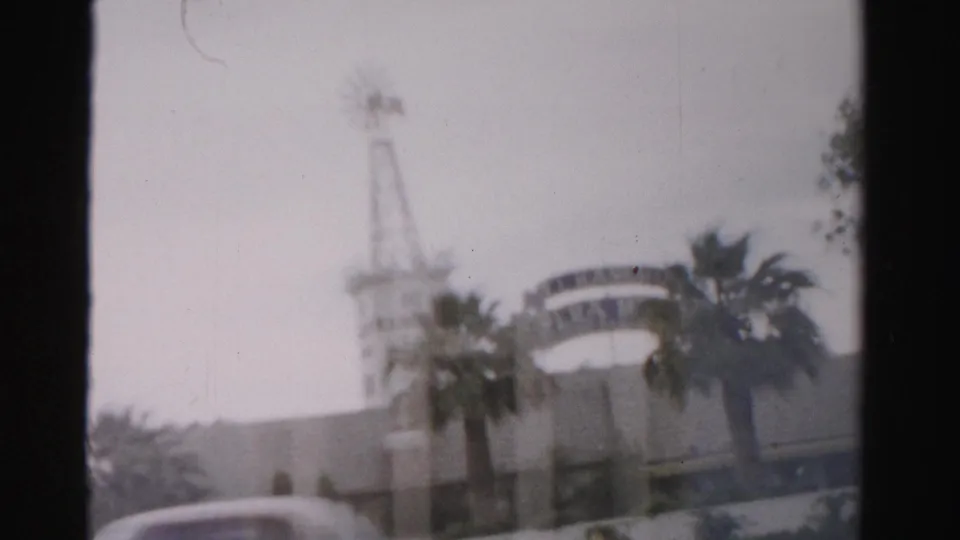 1957 Las Vegas frame showing a hazy exterior with a tower, curved sign-like structure, palms, low roofline, and partial vehicle foreground.