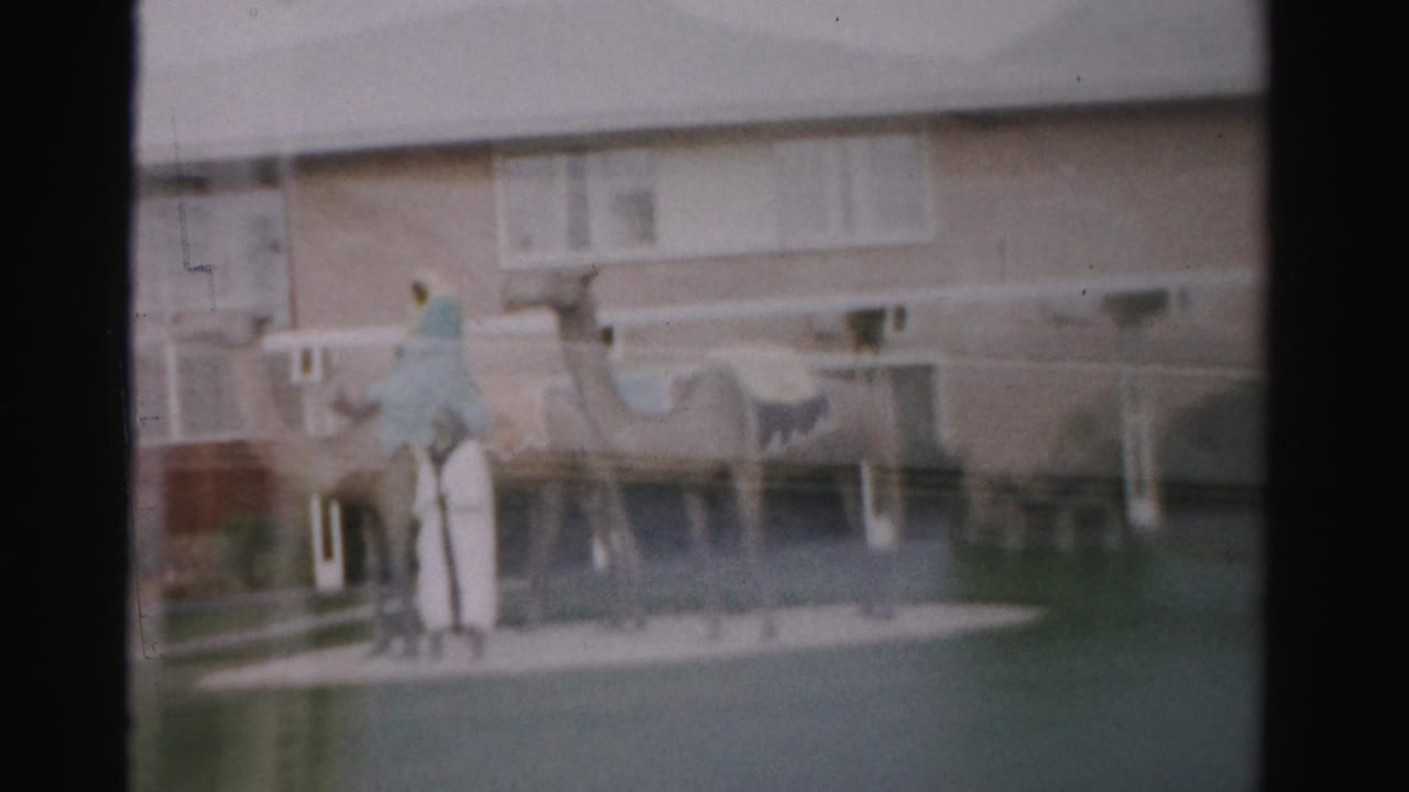 1957 Las Vegas home-movie frame showing two camels with a robed figure and a man near a low mid-century building.
