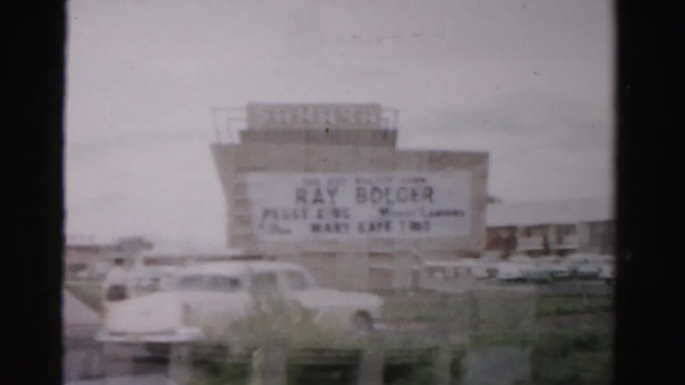 1957 Las Vegas home-movie frame showing a theater or drive-in marquee with readable performer names and parked cars nearby.